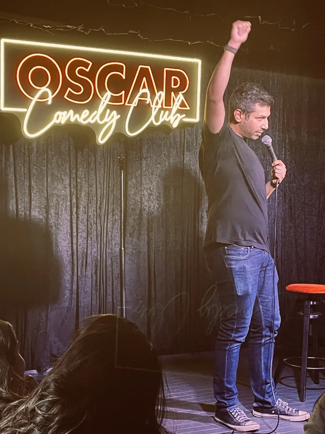 A comedian with his arm raised under a neon comedy club sign